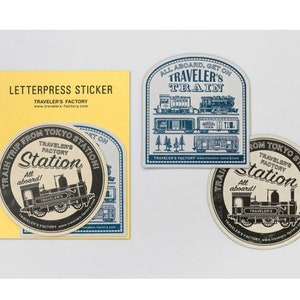May include: Three round letterpress stickers with a train theme. The stickers are cream coloured with black and blue lettering and illustrations. One sticker says "All aboard, get on Traveler's Train" and features a train with multiple carriages. The other two stickers say "Train trip from Tokyo Station! Traveler's Factory Station All aboard!" and feature a train engine.