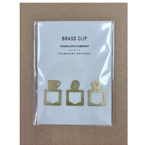 May include: A package of brass clips from Traveler's Company for Starbucks Reserve. The clips are gold-colored and feature a coffee cup, a leaf, and a flower design. The clips are packaged on a white card.