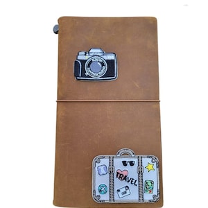 May include: A brown leather journal with a stitched closure and elastic band. The journal features two embroidered patches: a black and white camera and a suitcase with the word "TRAVEL" and travel-themed stickers.