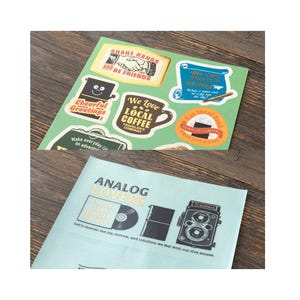 May include: A selection of stickers with diverse designs and text. The stickers display phrases such as "Share Hands and Be Friends," "We Love Local Coffee," and "Analog Lovers." The colour scheme includes green, yellow, and brown.