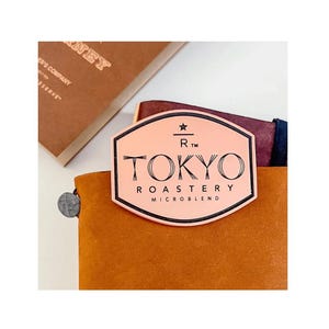 Op de afbeelding: Een roze en zwart Tokyo Roastery Microblend label op een bruin leren notitieboek. Op de achtergrond een bruin boek met gouden letters.