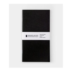 Könnte beinhalten: Schwarze Traveler's Factory Notizbuch-Nachfüllung mit schwarzem Papier. Das Notizbuch ist mit einem weißen Band umwickelt, das das Logo der Marke und Produktinformationen enthält. Das Notizbuch ist rechteckig und minimalistisch gestaltet.