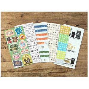 May include: Several sticker sheets with travel-themed designs, text, and patterns. The stickers feature a range of colours, including green, blue, orange, and yellow. They are arranged on a wooden surface, offering a variety of decorative options.