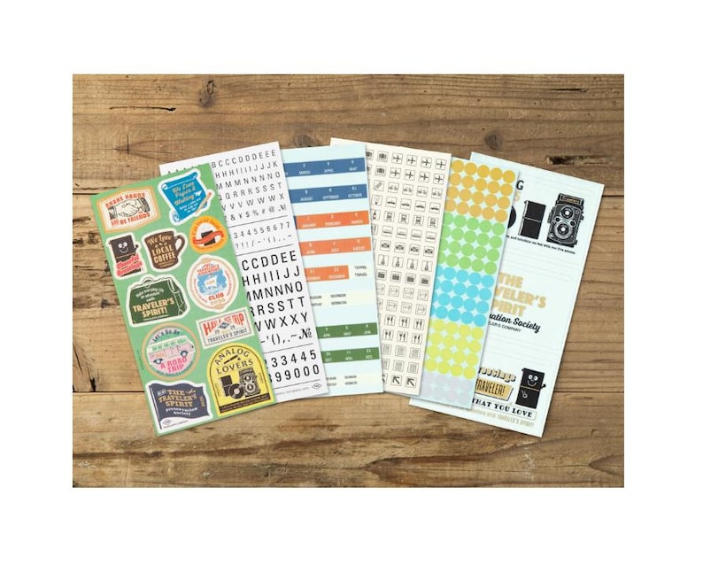 May include: Several sticker sheets with travel-themed designs, text, and patterns. The stickers feature a range of colours, including green, blue, orange, and yellow. They are arranged on a wooden surface, offering a variety of decorative options.