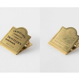Op de afbeelding: Twee goudkleurige metalen paperclips met een decoratief ontwerp. De eerste clip heeft de tekst "TRAVELER'S COMPANY JAPAN - TRAVELER'S notebook MADE IN JAPAN". De tweede clip heeft de tekst "Have a nice trip TRAVELER'S notebook TRAVELER'S COMPANY JAPAN".