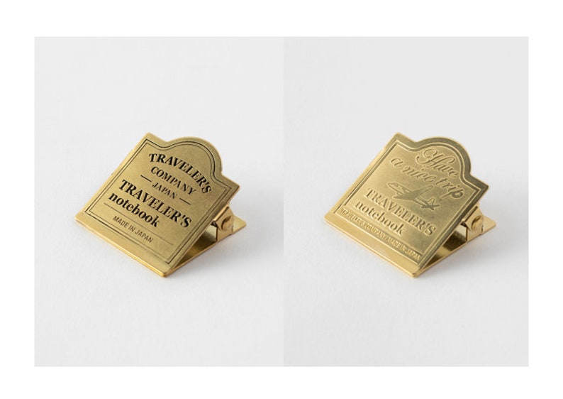 Op de afbeelding: Twee goudkleurige metalen paperclips met een decoratief ontwerp. De eerste clip heeft de tekst "TRAVELER'S COMPANY JAPAN - TRAVELER'S notebook MADE IN JAPAN". De tweede clip heeft de tekst "Have a nice trip TRAVELER'S notebook TRAVELER'S COMPANY JAPAN".