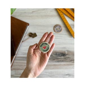 Puede incluir: Un parche bordado verde y blanco con un avión de papel rojo y el texto "TRUST THE PROCESS".