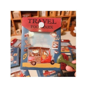 May include: A blue and red travel-themed pouch with the words "TRAVEL FOOD MARK" printed on the top. The pouch features illustrations of a tuk-tuk, camera, and other travel-related icons. A white button secures the pouch.