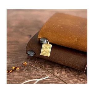 May include: Two leather journals, one brown and one dark brown, are stacked. A gold charm with the word "FRAGILE" is attached to the top journal. The charm features a design of two hands holding a plant. The journals are on a wooden surface.