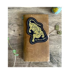 Peut inclure: Un journal en cuir marron avec un patch bleu marine représentant un tigre doré. Le patch porte l'inscription "The Superior Labor". Un petit globe et d'autres accessoires sont visibles en arrière-plan.