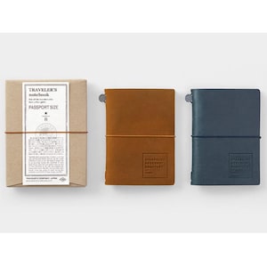 Op de afbeelding: Drie Traveler's Notebooks in paspoortformaat worden getoond. De set bevat een bruine papieren verpakking met tekst, een bruin leren notitieboek en een donkerblauw leren notitieboek. Elk notitieboek heeft een touwsluiting.