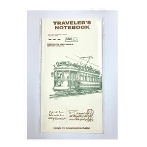 May include: A vintage-style traveller's notebook with a green and white illustration of a tram on the front cover. The cover also features the text "Traveler's Notebook" in gold lettering and a stamp that says "Mail Art".