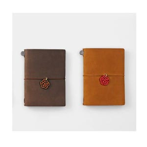 May include: Two leather-bound journals with a knotted string closure and a decorative knot charm. The journal on the left is brown and the journal on the right is orange.
