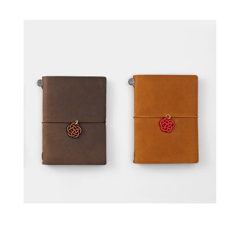 May include: Two leather-bound journals with a knotted string closure and a decorative knot charm. The journal on the left is brown and the journal on the right is orange.