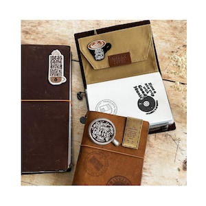 May include: Several brown leather journals and notebooks are displayed, adorned with coffee-themed patches and text. One journal features a patch that reads "Ideas come after coffee." Another has a coffee cup design and the text "Always coffee time."