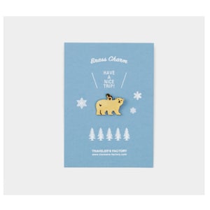 Op de afbeelding: Een messing beer-vormige bedel met een lus, gepresenteerd op een lichtblauwe kaart. De kaart bevat de tekst "Brass Charm" en "Have a Nice Trip!", plus illustraties van sneeuwvlokken en bomen. De bedel is ontworpen voor bevestiging.