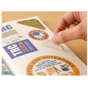 May include: A hand peeling a sticker from a notebook. The notebook is covered in travel-themed stickers, including ones that say "TRAVELER'S Cafe" and "TRAVELER'S AIRLINES". The stickers are in shades of brown, blue, and white.