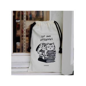 May include: A white drawstring bag with black drawstrings and a black graphic of a cat lying on a stack of books. The text "CAT AND NOTEBOOKS" is printed above the cat illustration.