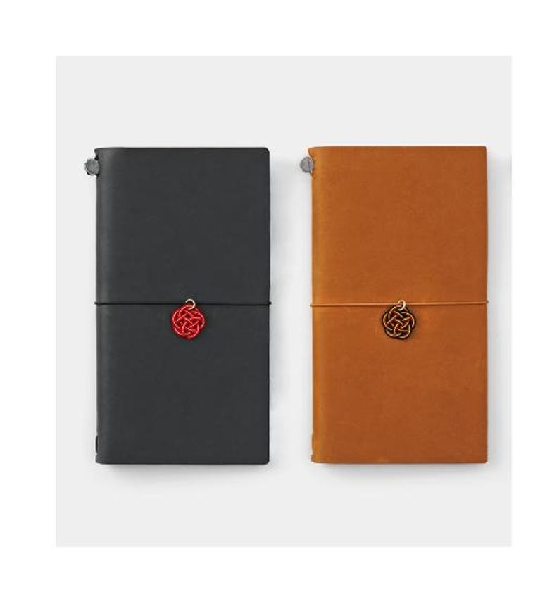 May include: Two leather-bound notebooks with a red and black string closure. Each notebook has a small red and black tassel charm.