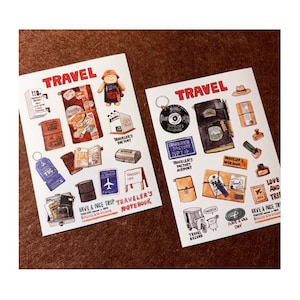 May include: Two white rectangular sticker sheets featuring colorful illustrations of travel-themed items, including notebooks, a bear, a record, and a tricycle. The text TRAVEL, TRAVELER'S FACTORY, TRAVELER'S NOTEBOOK, and HAVE A NICE TRIP is printed in red and black.