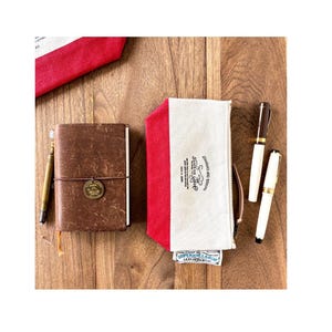 May include: A collection of stationery items on a wooden surface. Includes a brown leather journal, a red and white canvas pencil case with text, and two pens. The journal has a brass medallion and a pen attached. The pencil case has a zipper.