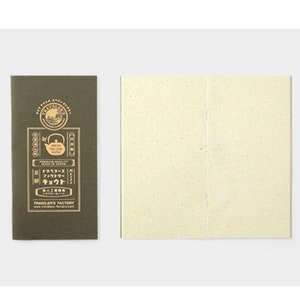 May include: A dark green paper sleeve with gold text and a cream-colored paper insert. The sleeve has Japanese text and a logo. The insert is a square of cream-colored paper with a vertical crease down the middle. The sleeve reads "Traveler's Factory."