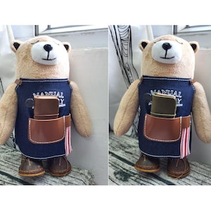 以下が含まれることがあります： デニムのエプロンを着たテディベアのぬいぐるみ。茶色の革のポケットと小さな茶色の革のポーチが付いています。エプロンには「MANUAL」と「Y」の文字がプリントされています。クマは茶色の革靴を履いています。