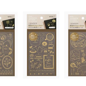 Puede incluir: Tres hojas de pegatinas con diseños de lámina dorada sobre un fondo marrón. Las pegatinas presentan varios temas, incluyendo motivos florales, de café y de viaje. Cada hoja tiene texto japonés y un pequeño gráfico de cuaderno negro.