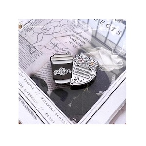 Peut inclure: Deux épingles en émail : une en forme de tasse à café avec le mot "coffee", et l'autre avec des fleurs et le texte "Mood Coffee Loading". Les épingles sont noires, blanches et argentées, sur un fond de planificateur.