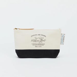 Könnte beinhalten: Eine Canvas-Tasche mit schwarzem Boden und cremefarbenem Oberteil. Die Tasche hat einen goldenen Reißverschluss und einen kleinen Leder-Zuglasche. Auf der Vorderseite ist der Text "STANDARD AND STRONG" und "THE SUPERIOR LABOR" aufgedruckt. Ein kleines Etikett an der Seite lautet "SUPERIOR LABOR LEATHER&CO."