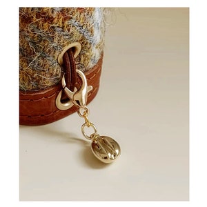 Può includere: Primo piano di una borsa in pelle marrone e lana con una chiusura in metallo dorato e un ciondolo a forma di chicco di caffè dorato.