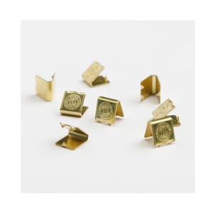 Könnte beinhalten: Eine Sammlung kleiner, goldfarbener Metall-Bandklemmen mit gefaltetem Design. Jede Klemme verfügt über ein kreisförmiges, geprägtes Logo auf der Vorderseite mit den Buchstaben TSL im Inneren sowie kleine Zinken zur Befestigung von Stoff oder Band.
