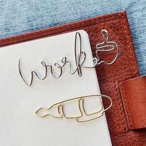 以下が含まれることがあります： 「work」の文字とコーヒーカップの形をした銀色のワイヤーのブックマークと、コーヒーカップの形をした金色のワイヤーのブックマークが白いノートの上に置かれています。ノートは茶色の革のジャーナルの中にあります。