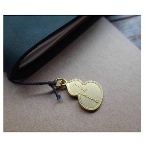 Op de afbeelding: Een goudkleurige metalen bedel in de vorm van een kalebas, bevestigd aan een donkerblauw koord. De bedel hangt aan een notitieboek. De bedel heeft Japanse tekst.