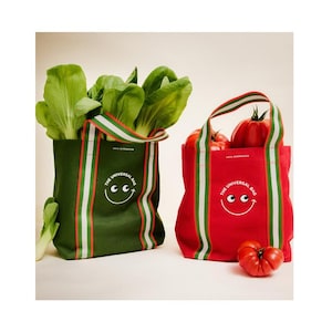Puede incluir: Un bolso tote verde oscuro lleno de bok choy y un bolso rojo lleno de tomates. Ambos bolsos tienen asas a rayas, un gráfico de cara sonriente blanca y el texto "THE UNIVERSAL BAG" impreso en blanco en la parte frontal de cada bolso.