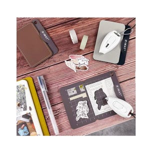 May include: A close-up of a craft tool with a white handle and a gray blade. The tool is on a gray cutting mat with a grid pattern. The mat is on a wooden surface with a sticker and two rolls of tape.