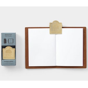 Op de afbeelding: Een messing clip met een gebouwontwerp is bevestigd aan een bruin leren notitieboek. Een kleine blauwe doos met de woorden "Brass Clip" en "Starbucks Reserve" staat naast het notitieboek. Het notitieboek is open op lege witte pagina's.