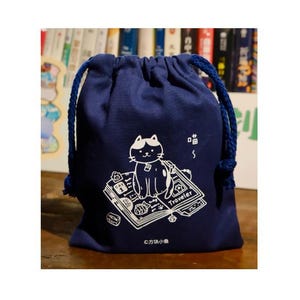 以下が含まれることがあります： 「Traveler」と書かれた本の上に座っている猫の白いイラストが特徴の、濃い青色の巾着袋。バッグには青いロープの引き紐と、猫の鳴き声の小さな白いグラフィックが付いています。「©方块小鱼」の文字が下部にあります。