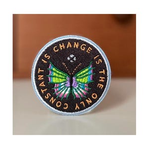 May include: Embroidered patch with a black background and a light blue border. The patch features a colorful butterfly with green, purple, and blue wings. The text "CONSTANT IS THE ONLY CHANGE" is in gold lettering around the edge.