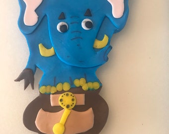May include: A blue fondant elephant with yellow tusks and brown eyes holding a brown pot with a yellow handle and gold accents. The elephant is on a stick.