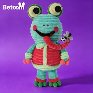May include: A plush frog toy in shades of teal, red, and green. The frog has large, round eyes, rosy cheeks, and a long red tongue with a small insect at the end. The toy is set against a purple background.