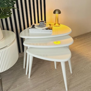 Puede incluir: Un conjunto de tres mesas nido blancas con tapas triangulares redondeadas y patas delgadas y anguladas. Una pequeña lámpara dorada y una taza con platillo están sobre la mesa superior. Las mesas están apiladas.