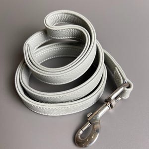 以下が含まれることがあります： 白のステッチが入った、丸まったライトグレーの革製犬用リード。 リードの端には銀色の金属製クラスプが付いています。 リードは柔らかく、質感のある素材で作られています。