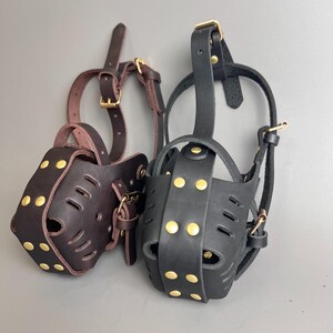 May include: Two leather dog muzzles, one dark brown and one black, are displayed against a gray background. Both muzzles have a basket design with ventilation holes, secured with straps and gold-colored metal hardware.