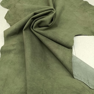 Puede incluir: Una pieza grande de cuero de ante verde oliva de forma irregular con una textura suave y mate. El material está drapeado en el centro, creando pliegues que revelan la parte inferior más clara, lisa y sin terminar de la piel.