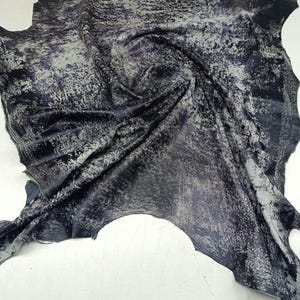 Può includere: Un pezzo di pelle con un colore di base blu navy scuro e una finitura metallica argentata screziata e usurata sulla superficie. Il materiale ha una forma organica irregolare con bordi grezzi e irregolari e un aspetto strutturato e piegato.