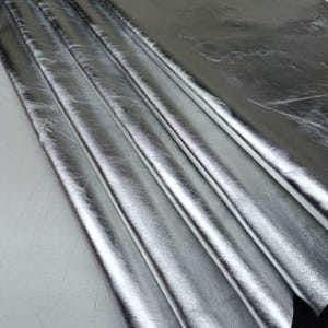 May include: A large, flexible sheet of metallic silver material with a reflective, high-gloss finish. The surface displays subtle natural creases and folds, creating deep shadows and bright highlights across the textured, shimmering expanse.