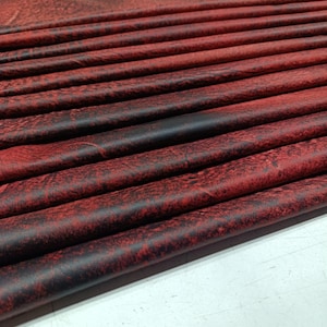 May include: Close-up of folded red and black leather material. The leather has a textured, distressed appearance with variations in color. The folds create a layered effect, showcasing the material's flexibility and depth. The material is likely used for crafting.