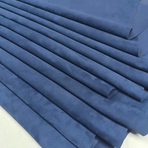 May include: A large, folded piece of deep blue suede leather. The material features a soft, matte, and velvety texture with visible natural grain variations across the surface. The fabric is draped in multiple layers, creating deep, smooth folds.