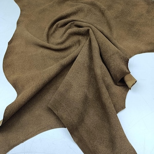 May include: A piece of brown suede leather with a soft, textured surface. The leather is folded and draped, showcasing its flexibility and natural shape. The color is a warm, earthy tone.